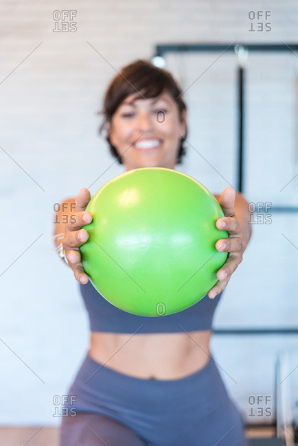 Soft focus of happy adult female exercising with green ball in outstretched arms during Pilates workout in gym
