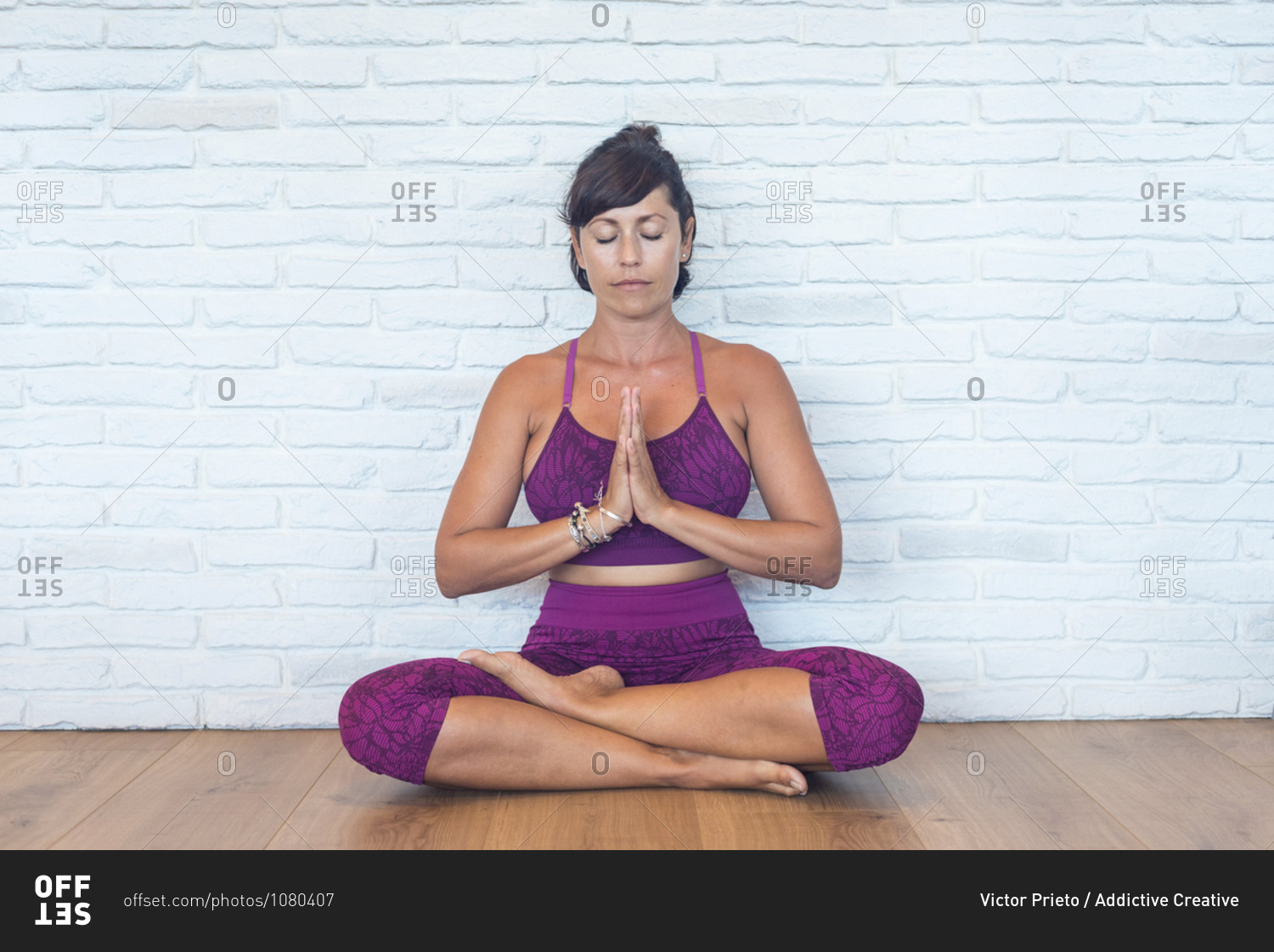 Adult female with closed eyes and clasped hands sitting in Padmasana pose meditating near brick wall during yoga lesson in studio