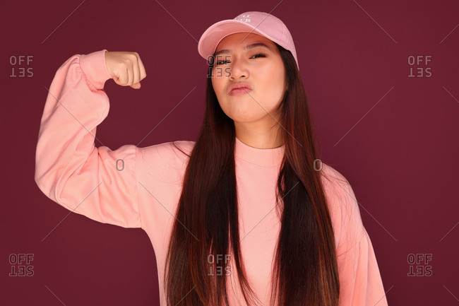 Portrait of funny young Asian woman in the studio looking at camera and showing biceps. She is wearing pink clothes over garnet background