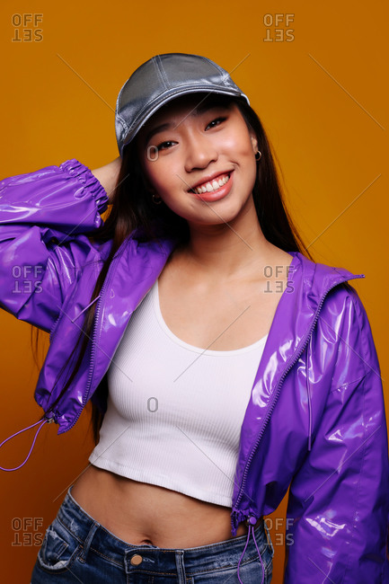 Portrait of happy Asian young woman. She wears a purple jacket and a grey cap and is looking at camera smiling against a yellow background