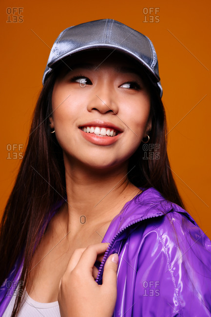 Portrait of happy Asian young woman. She wears a purple jacket and a grey cap and is looking away smiling against a yellow background
