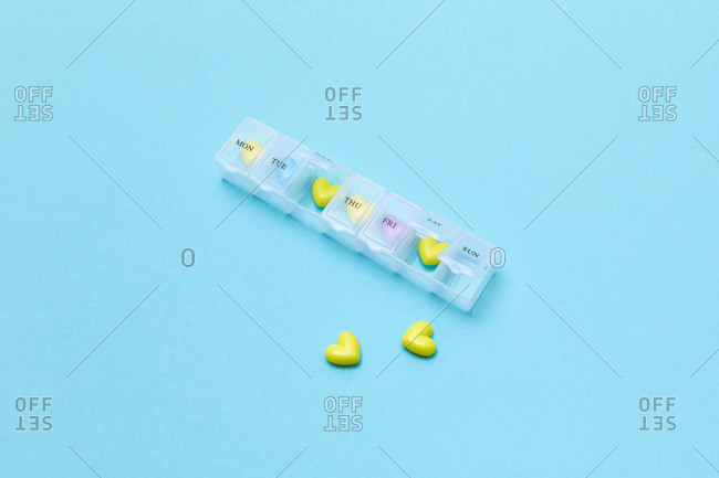 Scattered heart shaped pills and plastic container for week on colored blue background