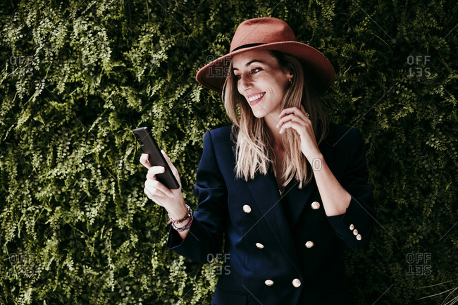 Happy fashionable woman using mobile phone against ivy