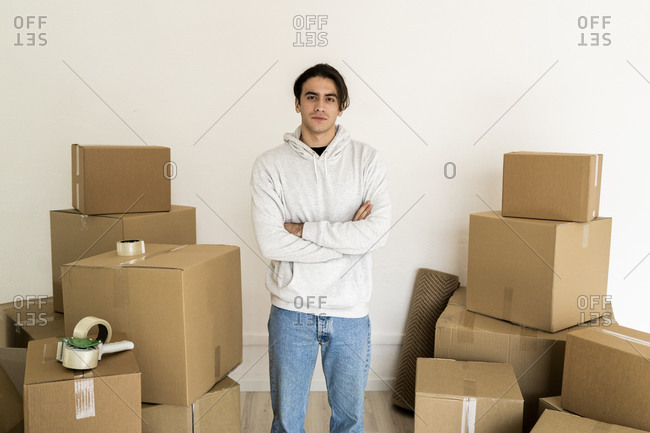 Young man with arms crossed standing amidst cardboard boxes in new house
