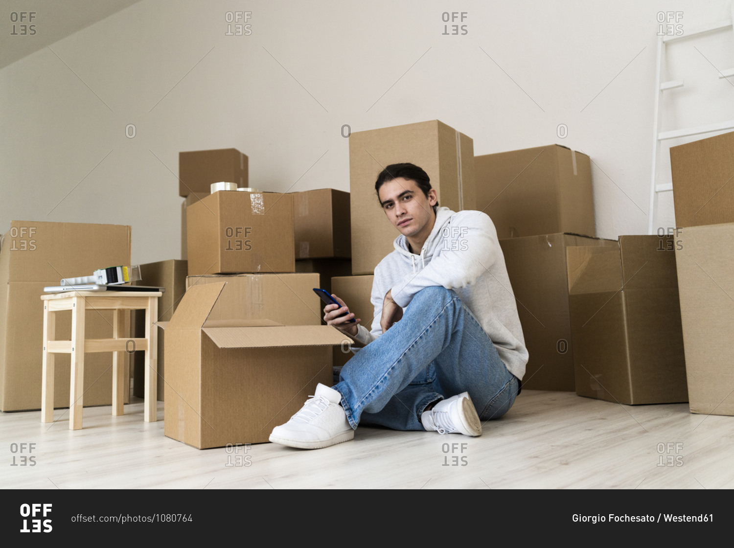 Young man using mobile phone while sitting against cardboard boxes in new house