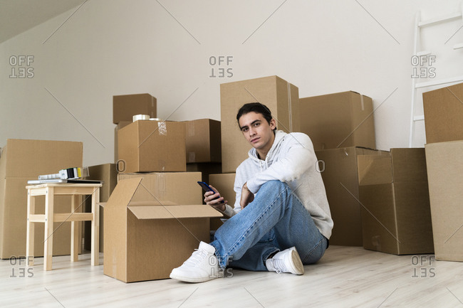 Young man using mobile phone while sitting against cardboard boxes in new house
