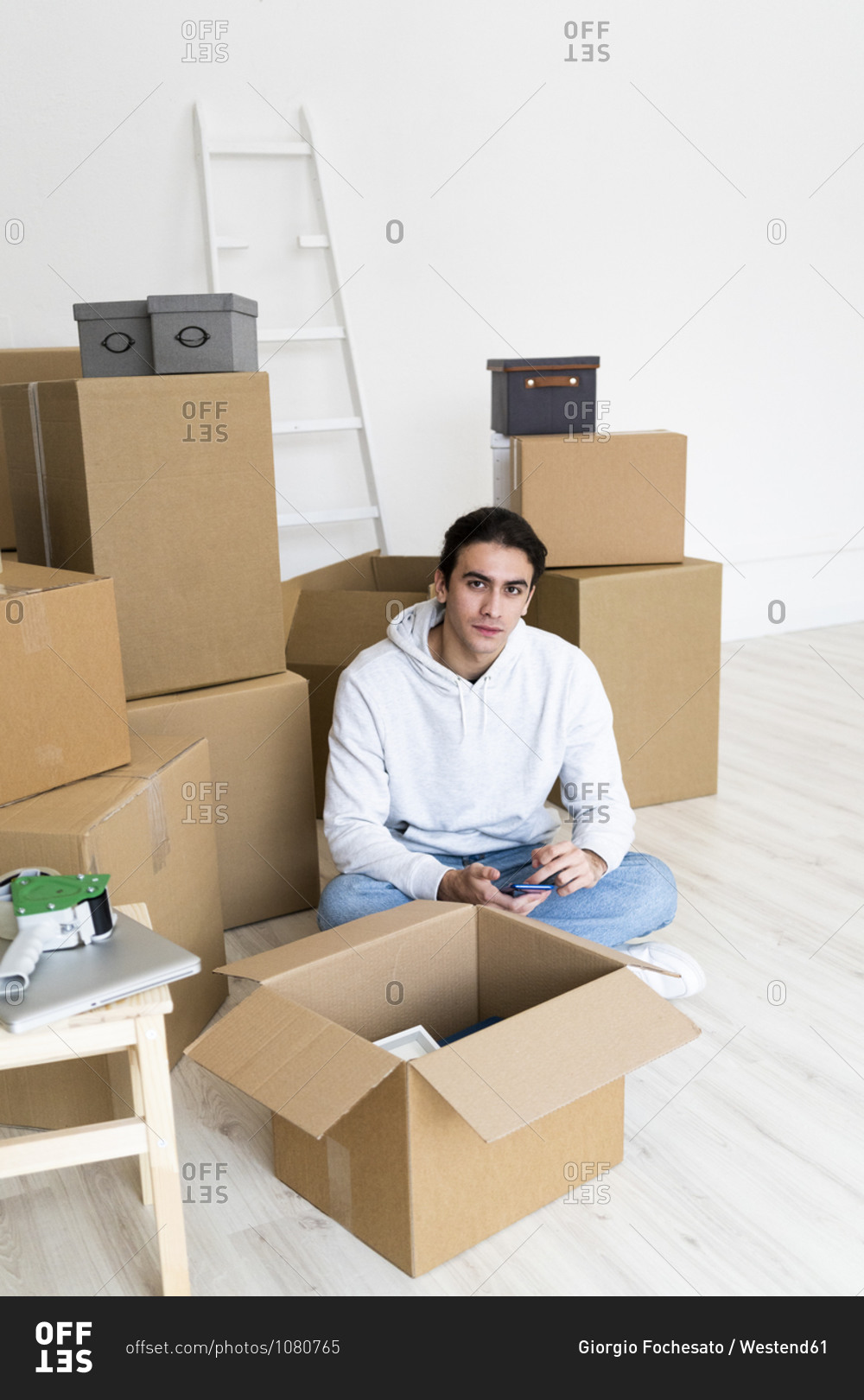 Young man with mobile phone sitting by cardboard boxes in new apartment