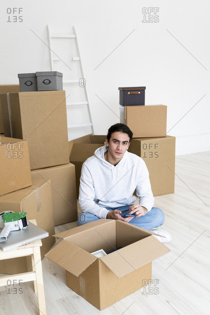 Young man with mobile phone sitting by cardboard boxes in new apartment