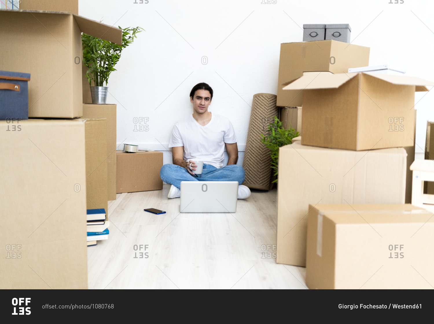 Young man drinking coffee while sitting on floor surrounded by boxes in new apartment