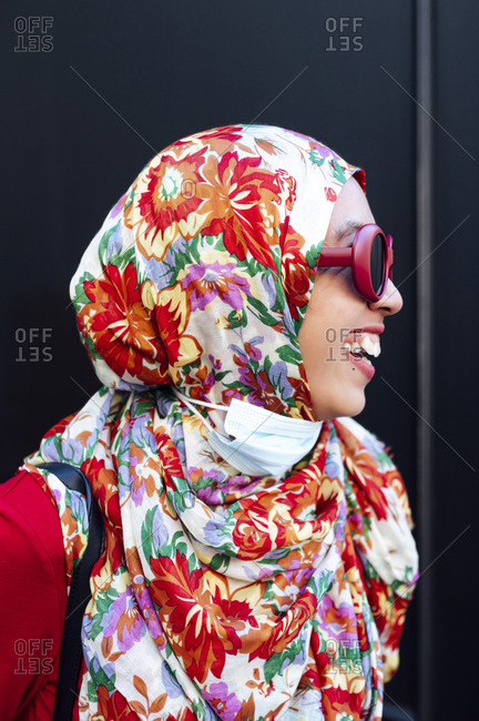 Cheerful woman in floral hijab against black wall during COVID-19
