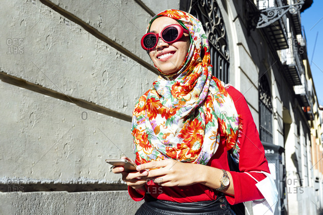 Smiling Muslim woman holding smart phone while walking by building on sunny day