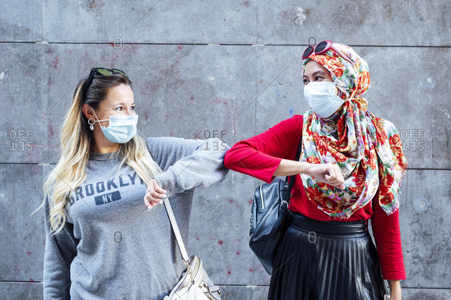 Female friends wearing protective face masks greeting with elbow bumps during COVID-19