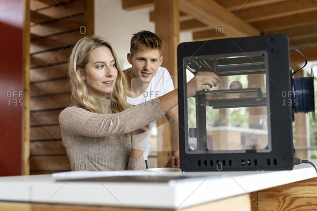 Businesswoman repairing 3D printer while sitting by colleague standing at home