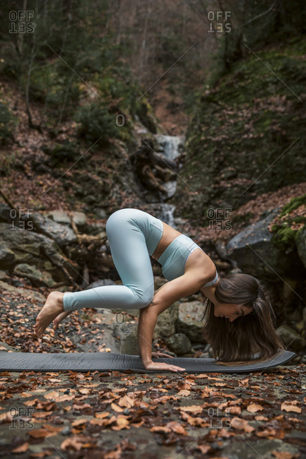 Athlete doing crow pose on exercise mat against waterfall in forest
