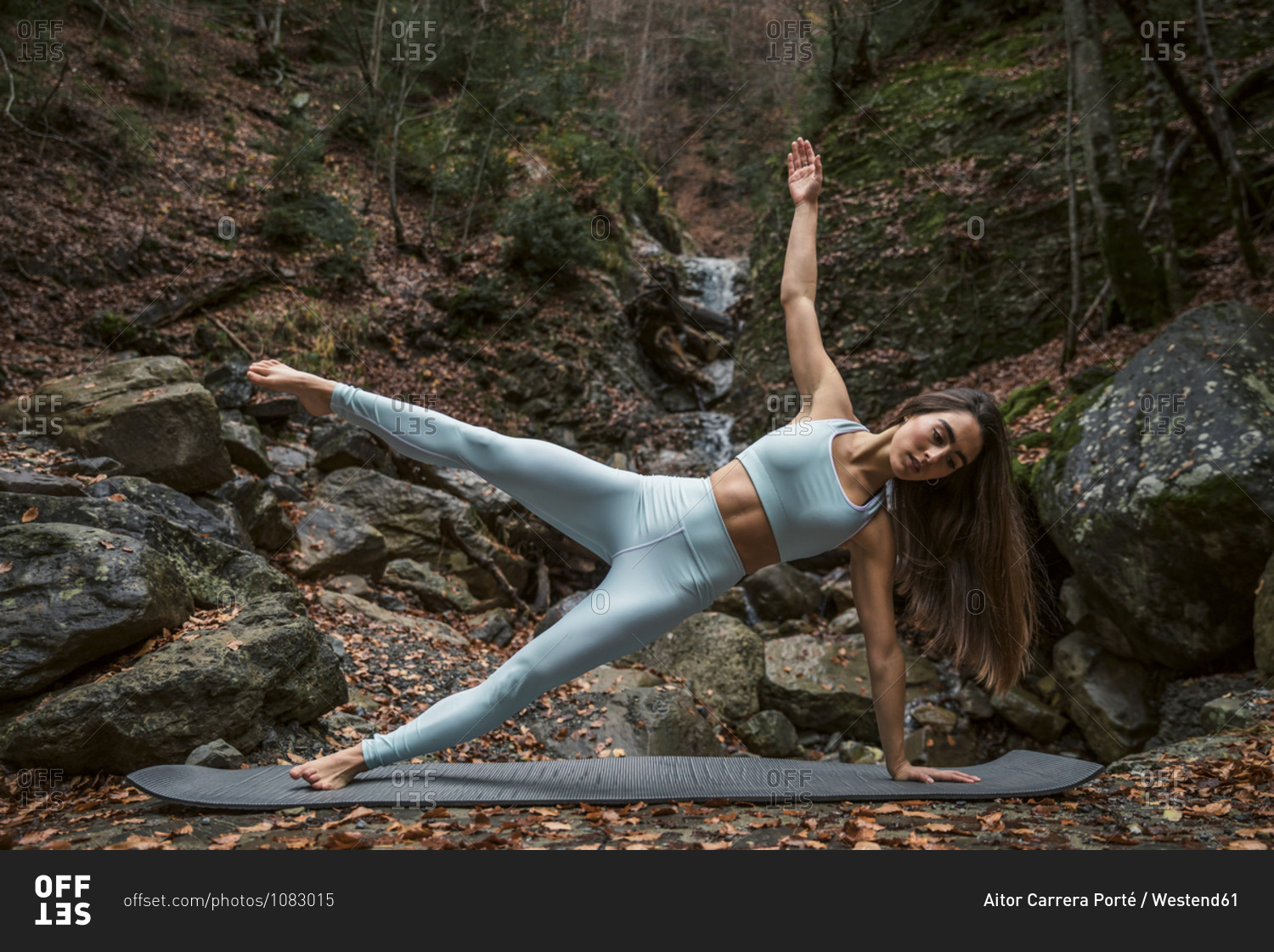 Young woman doing side plank pose on exercise mat against waterfall in forest