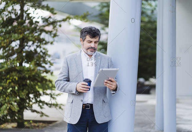 Male entrepreneur using digital tablet while holding coffee cup by column