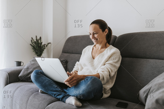 Smiling woman sitting cross-legged using laptop on sofa in living room