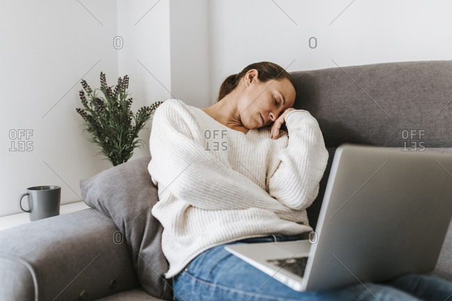 Tired woman sleeping while sitting with laptop on sofa in living room at home