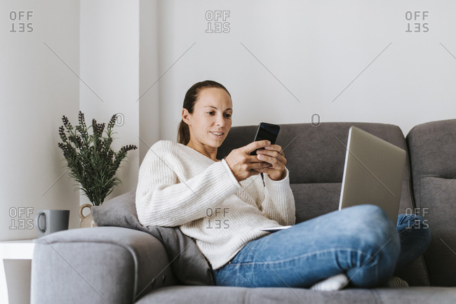 Woman text messaging through smart phone while sitting with laptop on sofa
