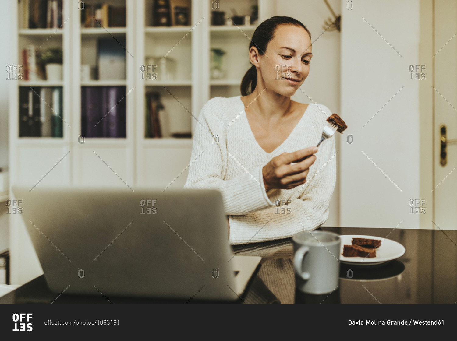 Woman eating chocolate brownie while sitting with coffee and laptop in illuminated kitchen