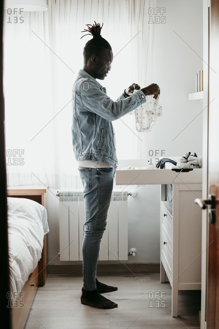 Young man holding baby clothes in bedroom at home