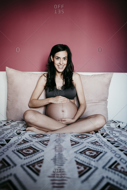 Smiling pregnant woman sitting on bed against red wall at home
