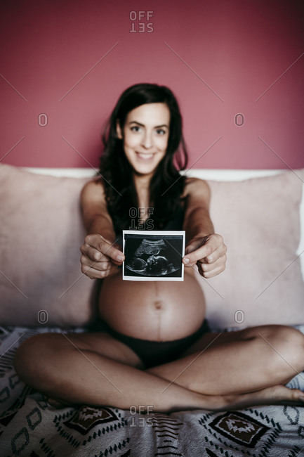 Mid adult pregnant woman showing ultrasound scan while sitting on bed at home