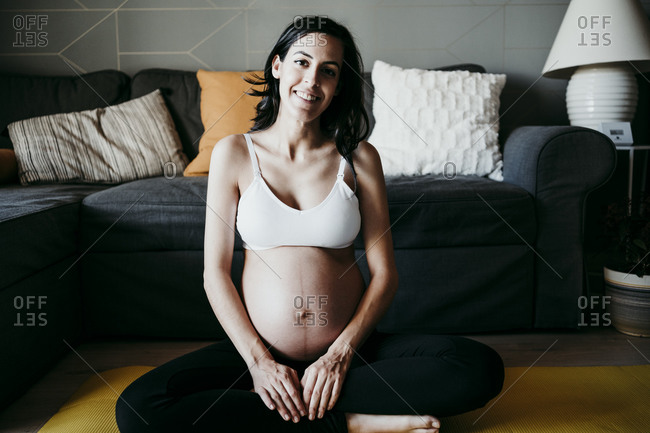 Smiling pregnant female in bra standing in white room and gently caressing  tummy while looking at camera stock photo - OFFSET