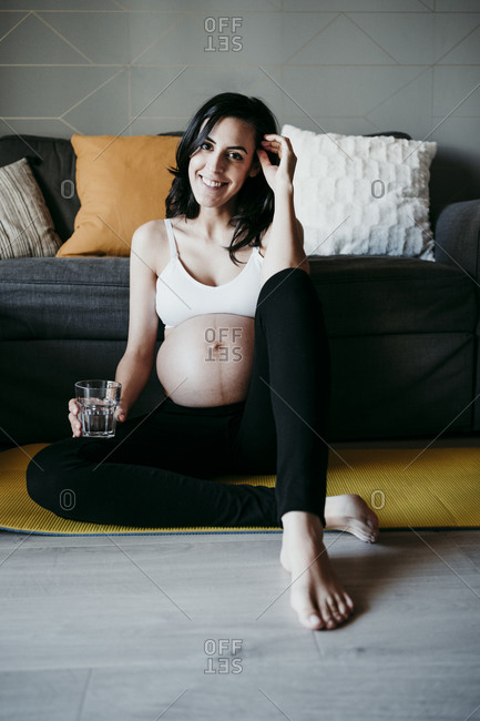 Smiling pregnant woman with glass of drinking water sitting on mat in living room at home