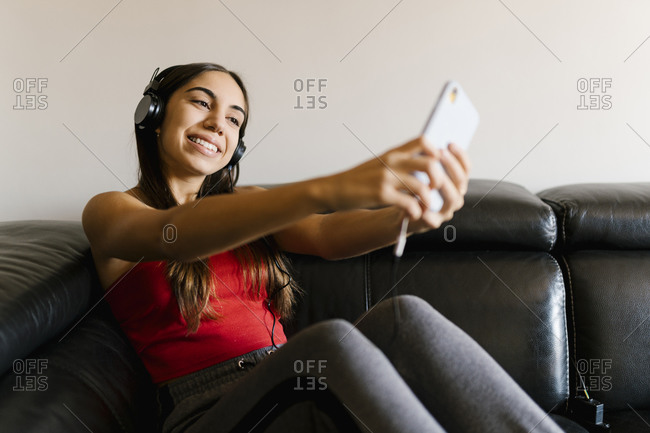 Teenage girl wearing headphones taking selfie through mobile phone while sitting on sofa at home