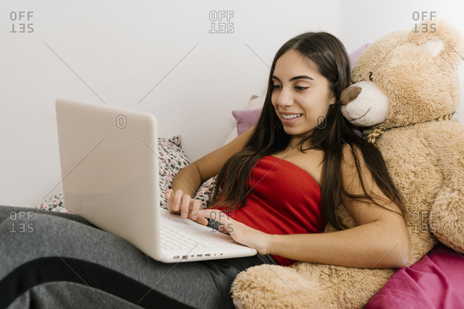 Smiling teenage girl using laptop while leaning on teddy bear toy at home