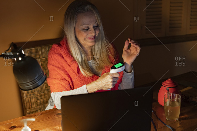 Smiling woman checking temperature with infrared thermometer while sitting with laptop at home