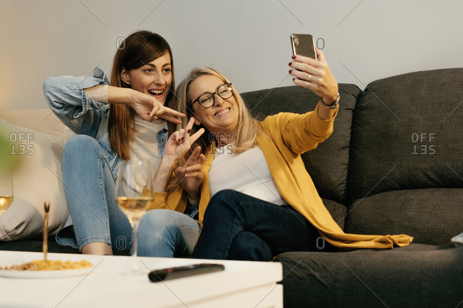 Smiling mother and daughter making peace sign while taking selfie through smart phone at home