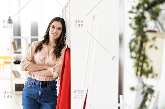 Smiling female fashion designer with arms crossed leaning on rack at studio