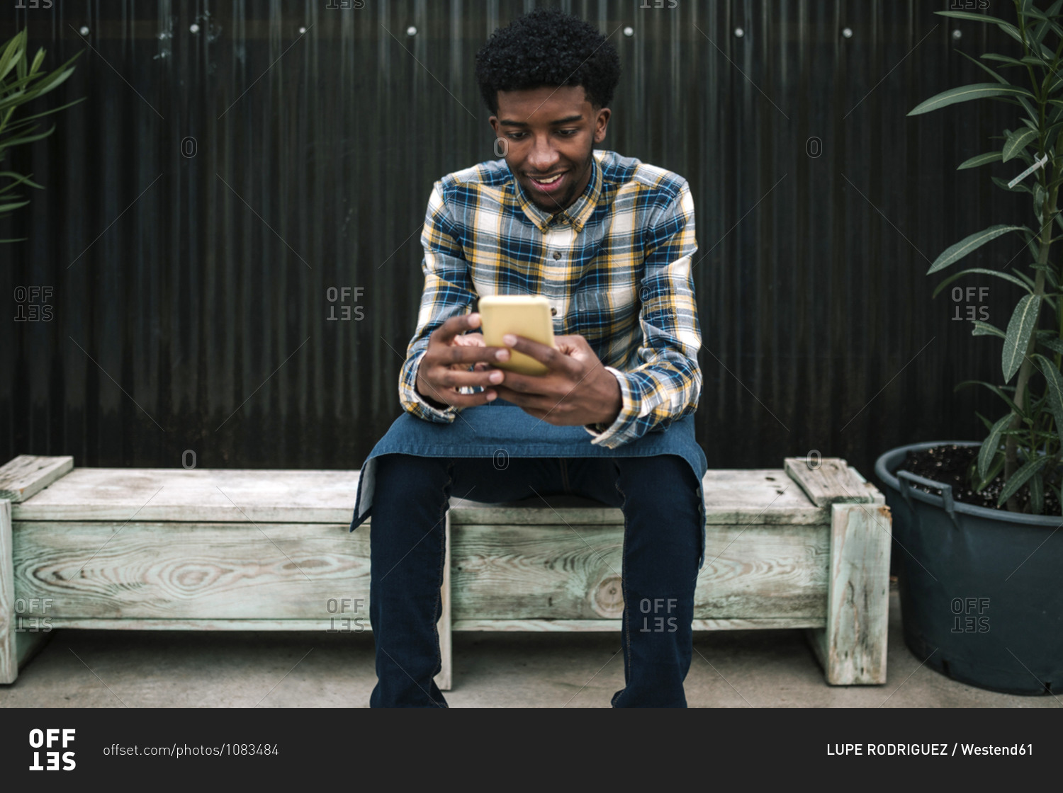 Smiling male farm worker using mobile phone while sitting on bench against corrugated wall