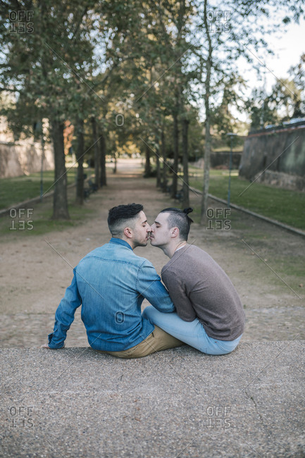 Affectionate gay couple kissing while sitting on footpath at public park