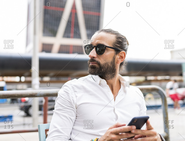 Portrait of bearded businessman wearing sunglasses sitting outdoors with smart phone in hands