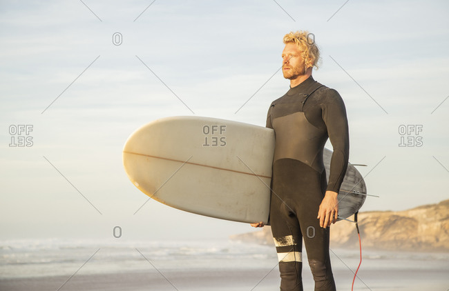 Blond man wearing wetsuit carrying surfboard while looking away at beach against sky