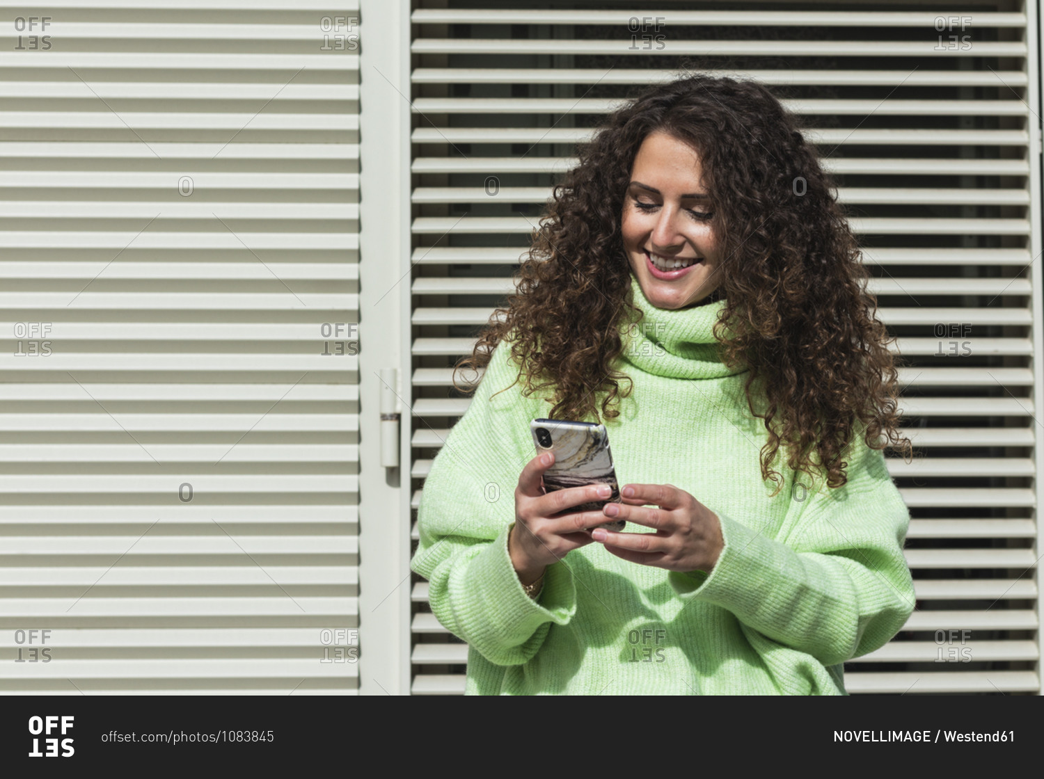 Smiling woman in neon green sweater using mobile phone against metallic door on sunny day