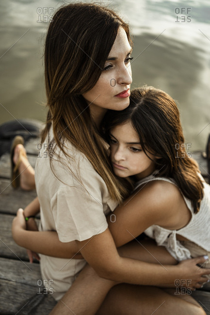Daughter embracing thoughtful mother while sitting on jetty