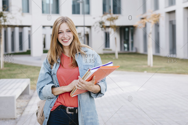 Smiling beautiful blond young woman holding books at university campus