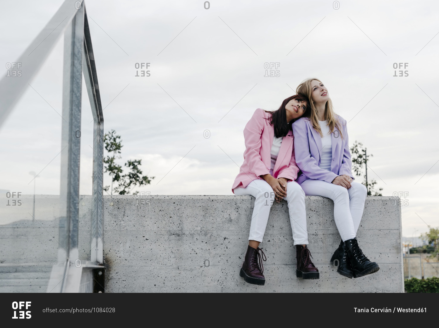 Young woman leaning on sister's shoulder while sitting on rooftop against  sky in city stock photo - OFFSET
