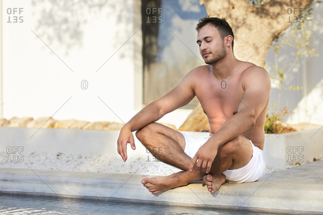Shirtless young man in shorts meditating at poolside