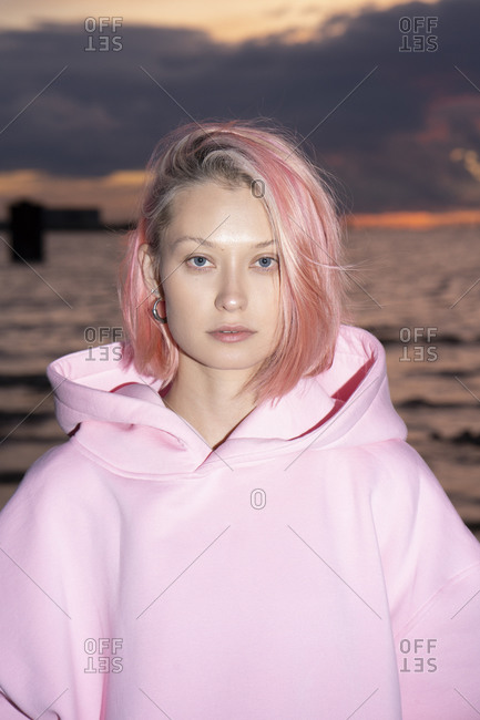 Portrait of young woman with pink hair wearing pink hooded shirt with sea at sunset in background