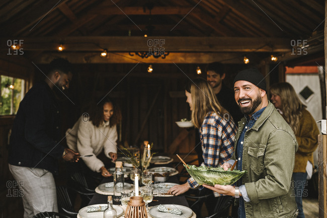 Portrait of smiling man holding leafy bowl while friends arranging dining table during party