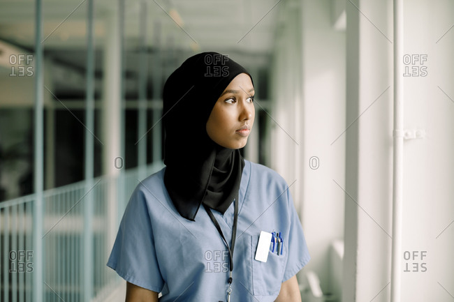 Contemplating female nurse looking through window while standing in hospital corridor