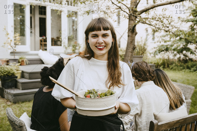 Portrait of smiling woman with food bowl standing in yard during social gathering