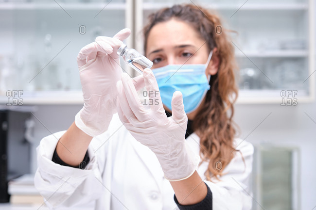 Young female scientist wearing face mask holding a coronavirus vaccine vial. Covid-19 vaccine development.
