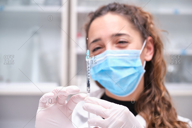 Young female scientist wearing face mask holding a coronavirus vaccine in a syringe and needle. Covid-19 vaccine development.