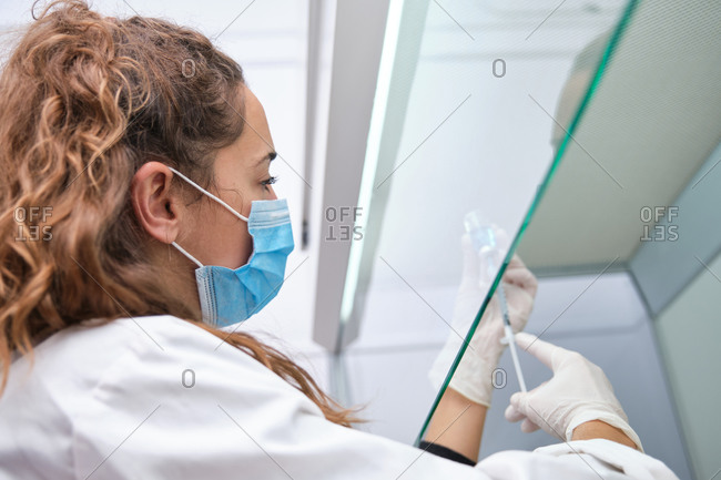 Young female scientist wearing face mask holding a coronavirus vaccine vial, syringe and needle in a laminar flow hood. Covid-19 vaccine development.